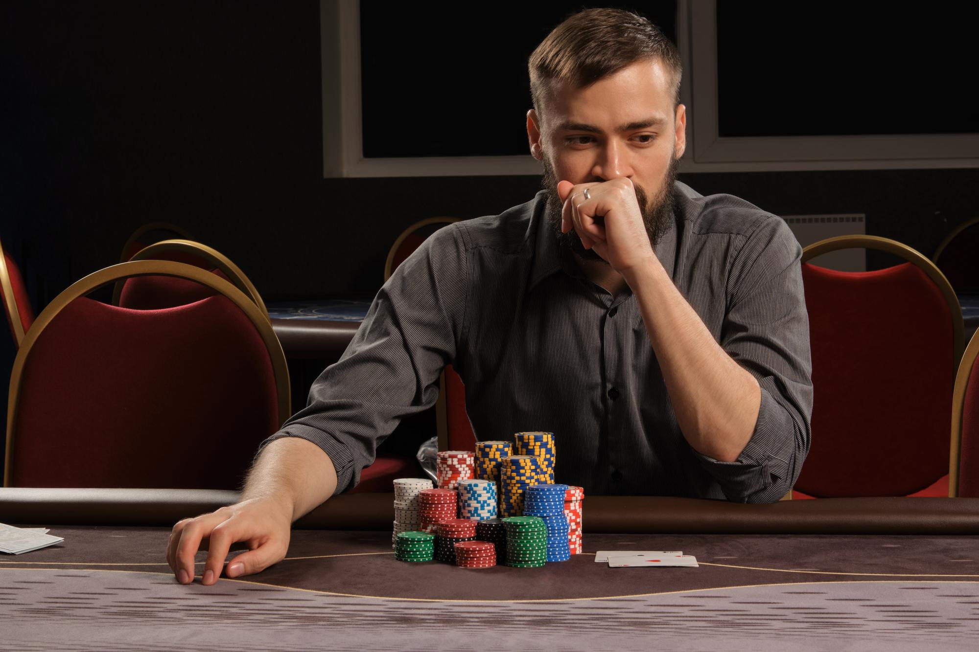 handsome-bearded-male-in-a-gray-shirt-is-playing-poker-sitting-at-the-table-in-casino-he-is-making-bets-waiting-for-a-big-win-gambling-for-money-games-of-fortune